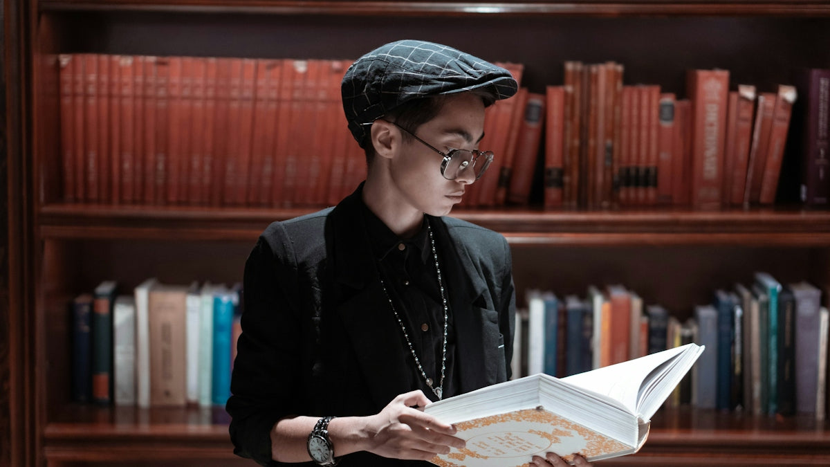 A young person reading a book in a library, wearing a black outfit and cap, showcasing stylish affordable women's party dresses and men's streetwear outfits.