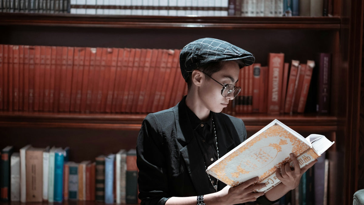 Person reading a book in front of a bookshelf, exploring latest arrivals women's men's fashion.