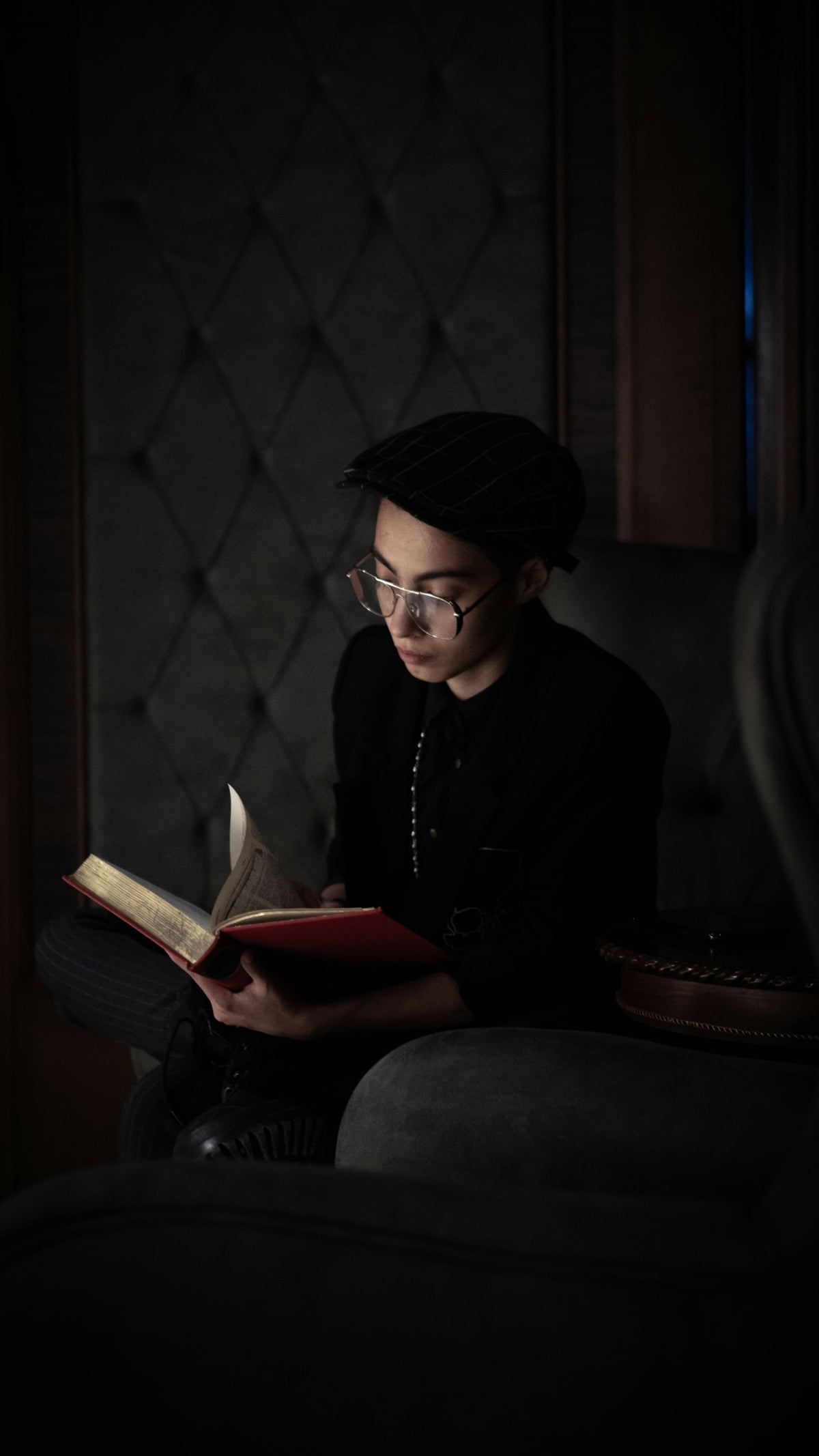 Young person reading a book in dim lighting, wearing glasses and a hat, absorbed in the text.