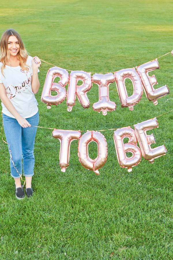 Rose gold balloon banner decor reading 'Bride To Be' held by a smiling woman in a park setting.